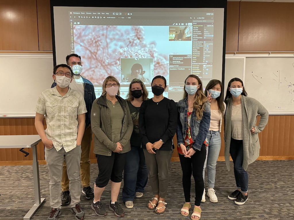 Eight teacher editors with masks on standing in front of a projected screen showing one additional teacher.